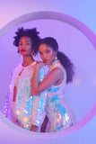  Two woman standing close together behind a circular port hole window wearing sparkly, sequin outfits by Rosa Bloom. 